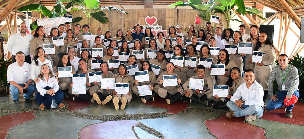 La imagen muestra a un grupo numeroso de personas, posiblemente una clase o un equipo, posando juntos para una foto de grupo. Todos están de pie delante de una estructura de madera, que podría ser un escenario o una pared. El grupo está formado por hombres y mujeres, y todos sostienen sus certificados