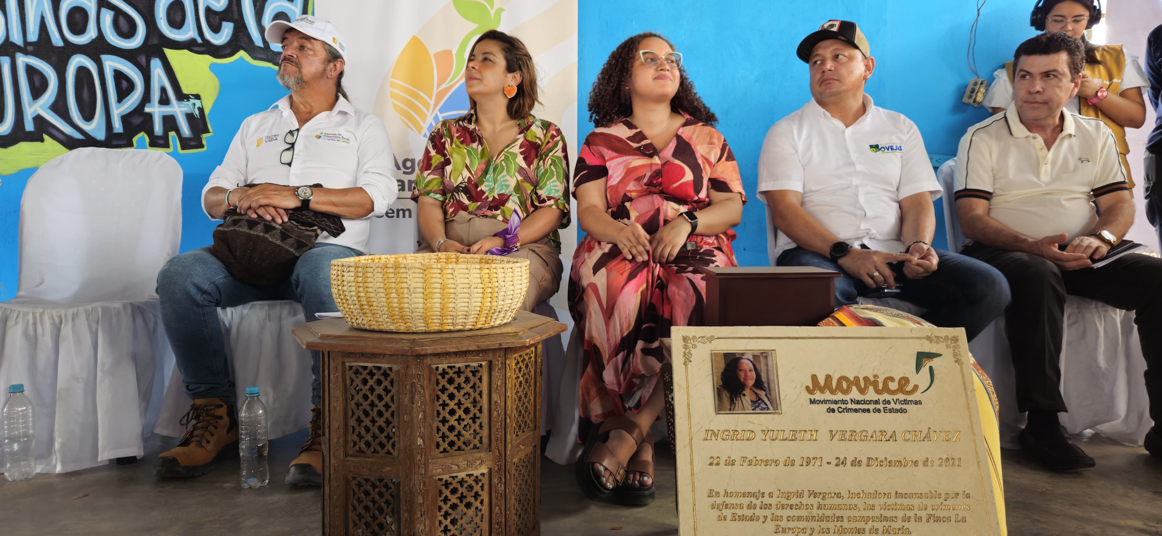 La imagen muestra a un grupo de personas sentadas en sillas, probablemente en un evento o reunión. Parecen ir vestidas con ropa informal, y algunas llevan sombreros y gafas de sol, lo que sugiere que podría ser un día soleado. Hay botellas en el suelo, posiblemente de agua u otras bebidas, lo que indica que el acto se celebra al aire libre o en un lugar donde es necesario hidratarse.  En primer plano, hay un cartel con una foto y algo de texto, que parece estar relacionado con el evento o con las personas presentes. El texto no es totalmente legible, pero incluye el nombre "Mónica" y menciona "Europa", lo que podría ser una referencia a un contexto europeo o a un nombre. El cartel también incluye una fecha, "22 de Febrero de 2017", que indica que el evento tuvo lugar el 22 de febrero de 2017.