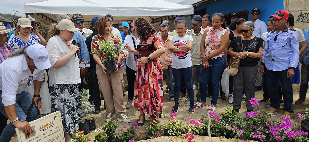 En la imagen, hay un grupo de personas reunidas en torno a la ministra de agricultura Jhenifer Mojica que sostiene una maceta. El escenario parece estar al aire libre, posiblemente en un evento o reunión de la comunidad, como indica la presencia de tiendas de campaña y plantas en macetas.