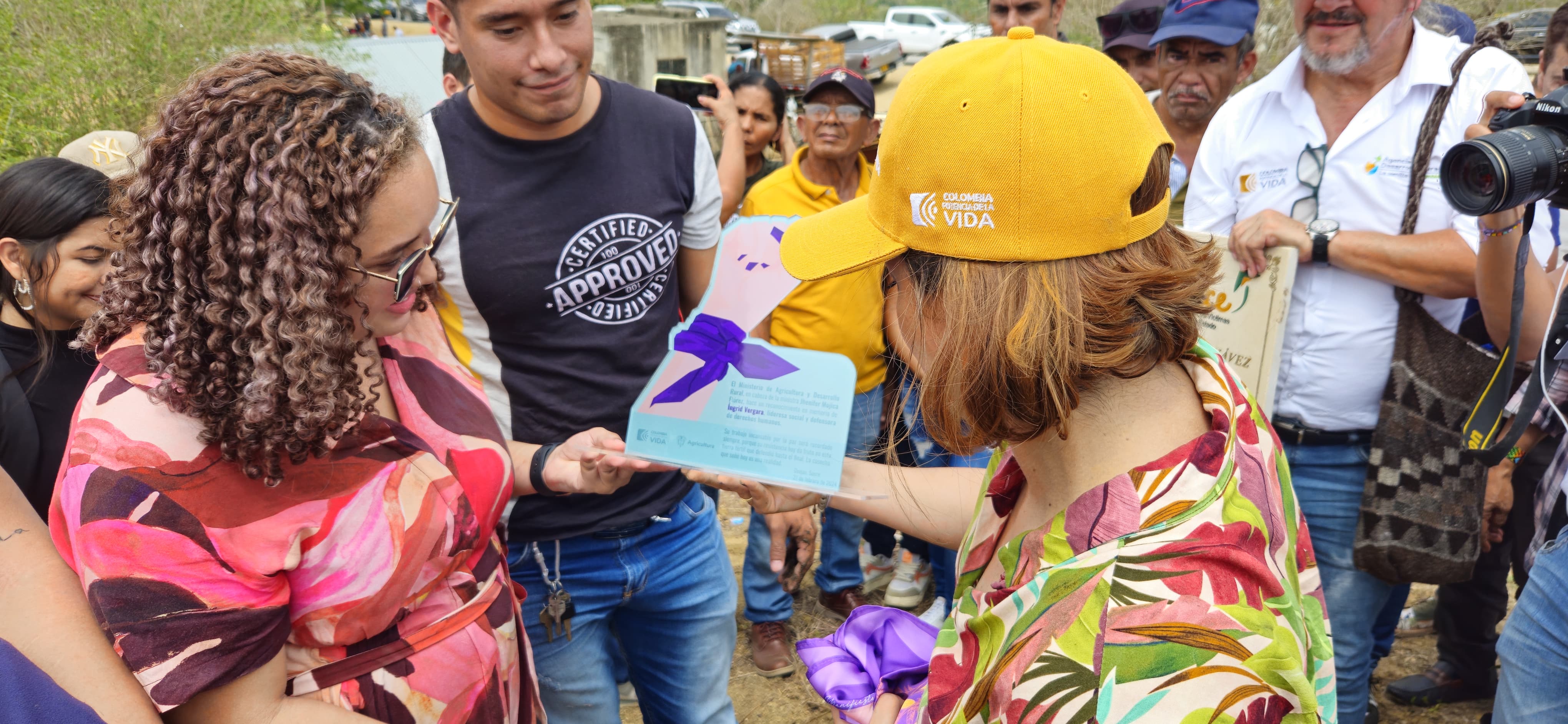 En la imagen hay dos personas que parecen mantener una conversación o un intercambio. La persona de la izquierda sostiene un papel o una tarjeta, que parece ser el centro de su interacción. La persona de la derecha lleva una gorra amarilla y mira el papel o la tarjeta con interés. Ambos sonríen, lo que sugiere una interacción positiva.