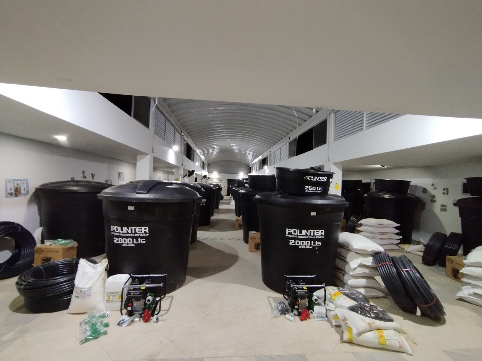 La imagen muestra un espacio interior, posiblemente un almacén o un área de almacenamiento, con un gran número de contenedores de plástico negro. Estos contenedores llevan la etiqueta «POULTRY 2000 L», lo que indica que probablemente están destinados a la industria avícola y tienen una capacidad de 2000 litros.