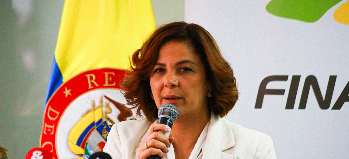 La imagen muestra a la ministra Jhenifer Mojica hablando por un micrófono. Parece estar en un acto formal, como indica la presencia de una bandera y un logotipo en el fondo.
