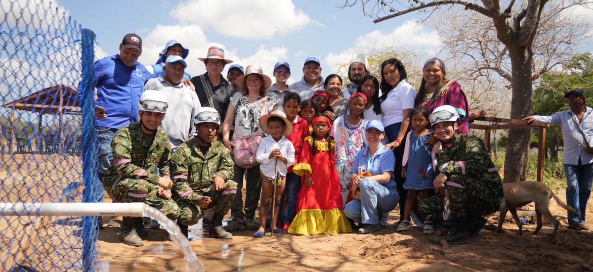 En la imagen, hay un grupo de personas reunidas al aire libre, posiblemente en un entorno rural o semirural. El grupo está formado por adultos y niños, y se encuentran de pie frente a una valla azul. Algunas personas llevan uniformes de tipo militar, lo que sugiere que podrían formar parte de una fuerza militar o de seguridad. El resto del grupo viste ropa informal y hay una mezcla de personas de diferentes orígenes étnicos.