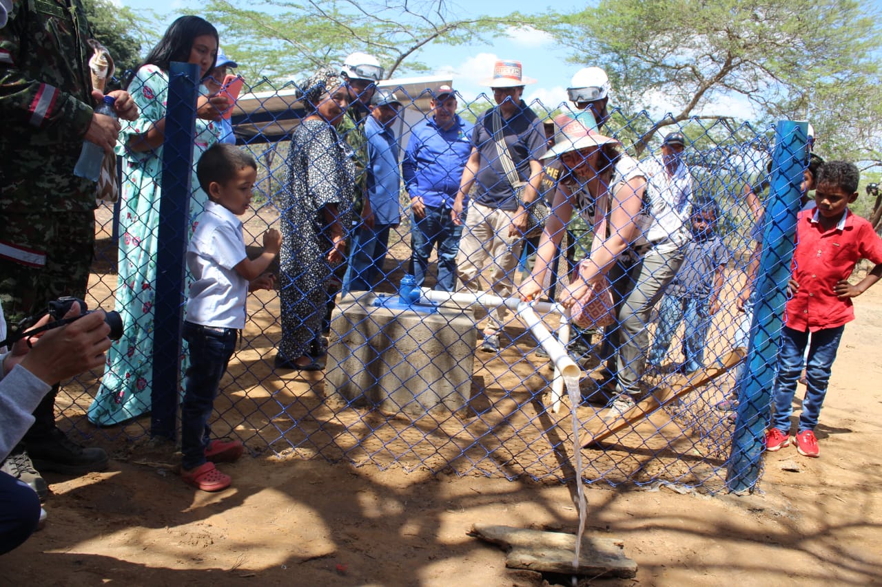 En la imagen, hay un grupo de personas reunidas en torno a un pozo de agua o una instalación de bombeo de agua. El escenario parece estar al aire libre, posiblemente en una zona rural o semirrural, dada la presencia de una valla y el entorno natural. Las personas están vestidas con ropa informal y algunas llevan sombrero, lo que sugiere que podría ser un día soleado.  El centro de la reunión parece ser la bomba de agua o el pozo, que está siendo manejado por una persona. El agua se bombea a un recipiente o depósito y la gente observa el proceso. Podría tratarse de una demostración, un acto comunitario o una actividad práctica relacionada con el acceso al agua y el saneamiento.