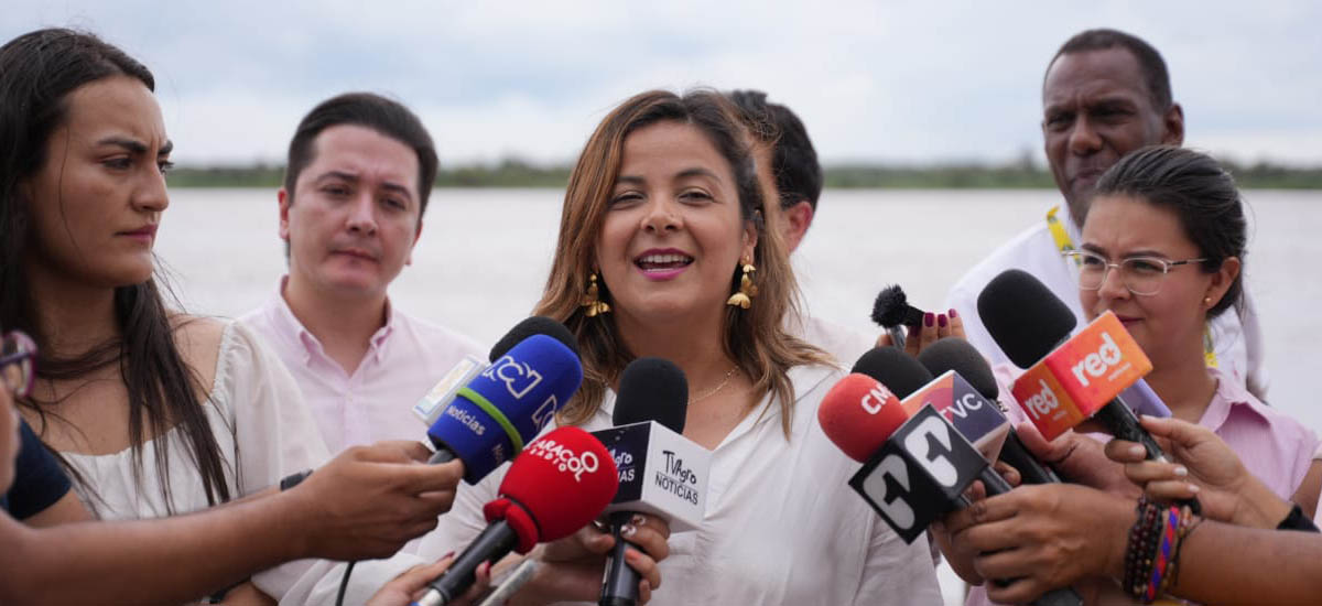 En la imagen se puede observar, a la Ministra Jhenifer Mojica, se ve que esta dando una rueda de prensa, de fondo se ve un paisaje con lo que parece ser un rio o lago y un cielo claro.