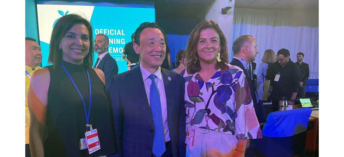 En la imagen se puede ver a la Ministra Jhenifer Mojica, junto con dos personas más posando para la foto, de fondo se puede ver una pantalla con un logo, en lo que parece una reunión 