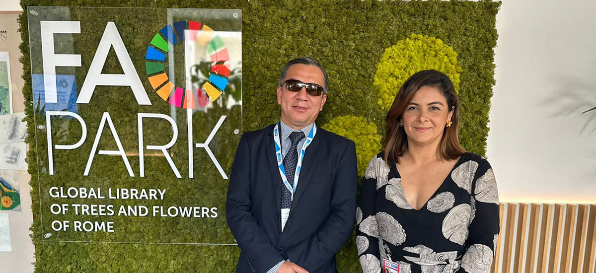 En la imagen se puede ver a la Ministra Jhenifer Mojica, posando junto a un señor, con un fondo verde, que parece ser pasto sintético, junto con un logo.