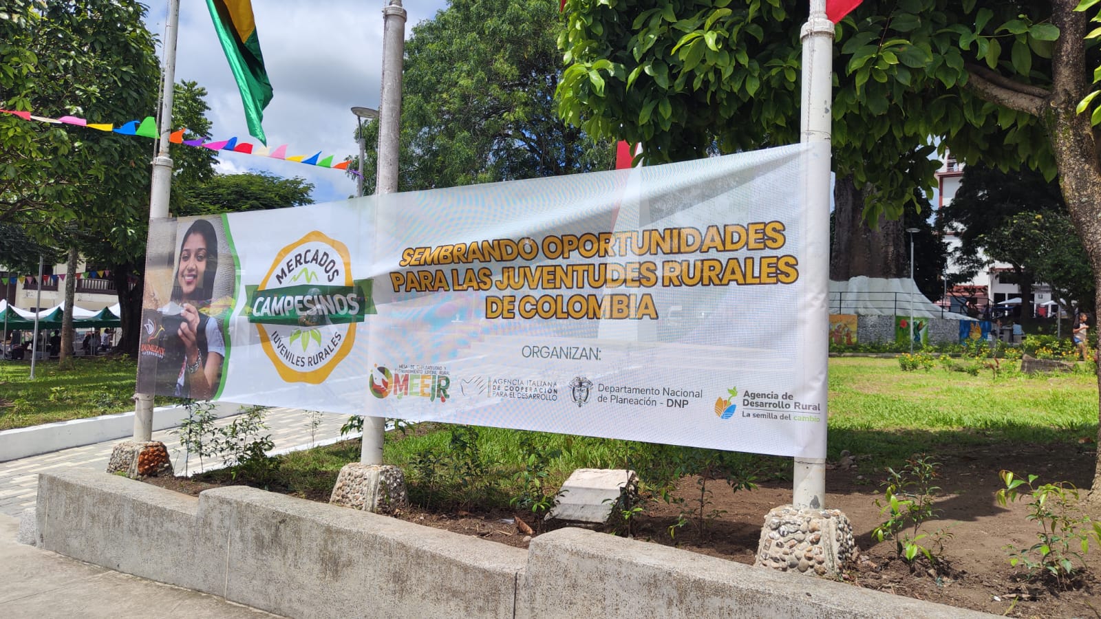 La imagen muestra una gran pancarta colgada de postes en lo que parece ser un parque o espacio público. tiene un mensaje que dice  «Creando oportunidades para la juventud rural en Colombia». También menciona a la Organización de las Naciones Unidas y al Departamento Nacional de Planeación de Colombia.
