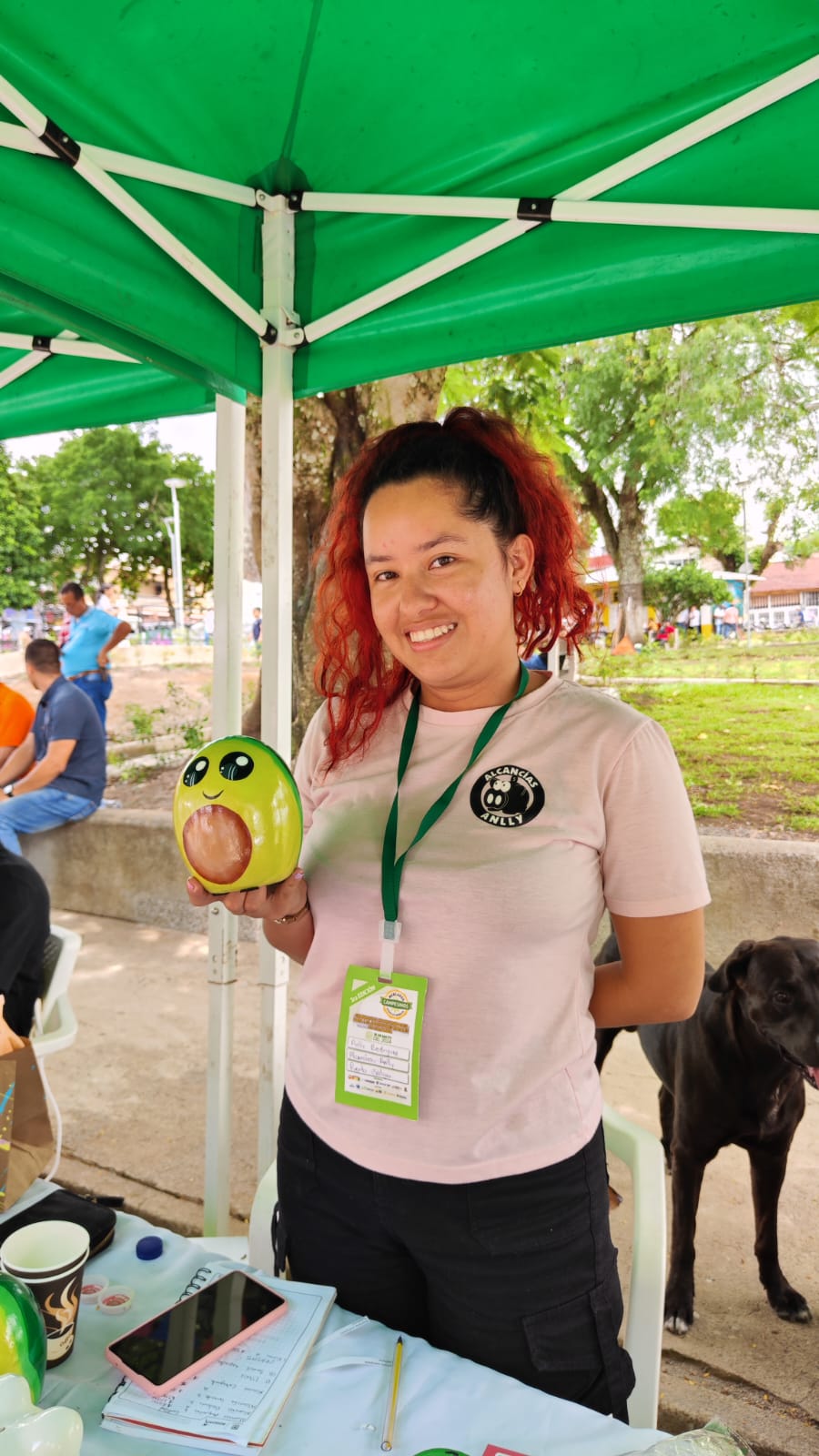 En la imagen, hay una persona de pie bajo una tienda verde. La persona sonríe y sostiene un objeto verde que parece ser un juguete o un elemento decorativo. Lleva una camiseta rosa con un logotipo en la parte delantera y un cordón verde alrededor del cuello, lo que sugiere que podría ser un empleado o un voluntario de un evento.