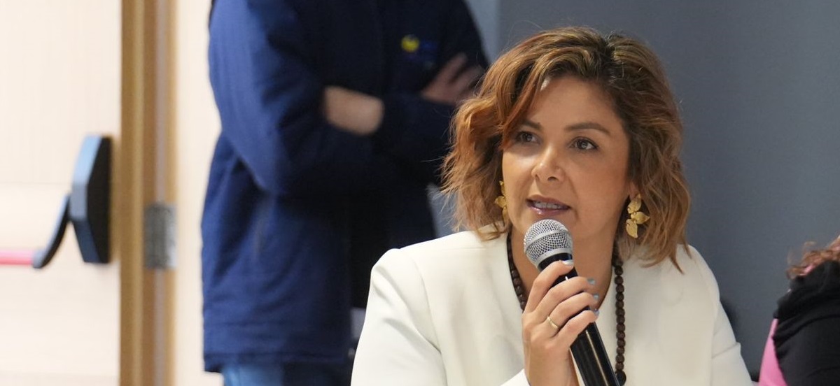 En la imagen se puede apreciar a la ministra de Agricultura Jhenifer Mojica, la cual se encuentra hablando por medio de un micrófono, la ministra tiene puesto un traje blanco muy elegante.
