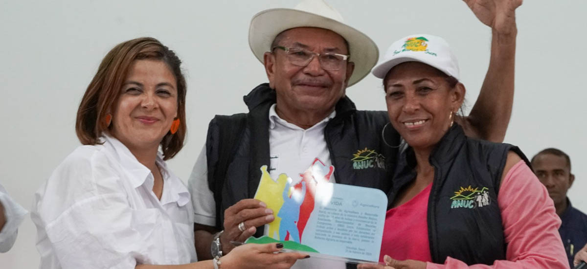 En la imagen aparecen tres personas posando juntos.  A la izquierda, aparece la ministra de agricultura Jhenifer Mojica, con blusa blanca, sonriendo y mirando a la cámara. En el centro, un hombre mayor con gafas, sombrero blanco y chaleco oscuro sobre camisa blanca. Sostiene un papel o un certificado con ilustraciones de colores. A la derecha hay otra mujer con un top rosa, un chaleco negro y una gorra blanca con un logotipo. 