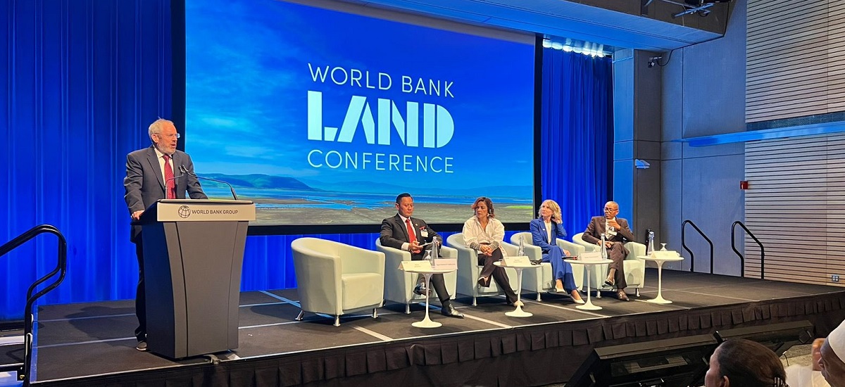 La imagen muestra una mesa redonda en una conferencia. Hay cinco personas en el escenario, una de ellas de pie en un podio, presumiblemente moderando o hablando, y otras cuatro sentadas en fila, probablemente panelistas u oradores. En el telón de fondo aparece el logotipo del Banco Mundial y la palabra «LAND» (tierra) en un lugar destacado, lo que sugiere que la conferencia está relacionada con cuestiones sobre la tierra, que podrían incluir temas como el uso de la tierra, los derechos sobre la tierra, la gestión de la tierra o la política de tierras.