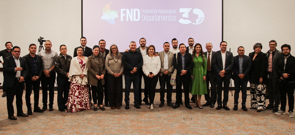 Imagen de un grupo de personas posando para una fotografía. Parecen estar en un evento formal o una conferencia, como indica el telón de fondo con el texto «FND Departamentos 30» y el logotipo de la «Federación Nacional de Departamentos». Las personas están vestidas con traje de negocios, algunas con corbata, y están de pie en una fila, que es una disposición común para las fotos de grupo en eventos profesionales.