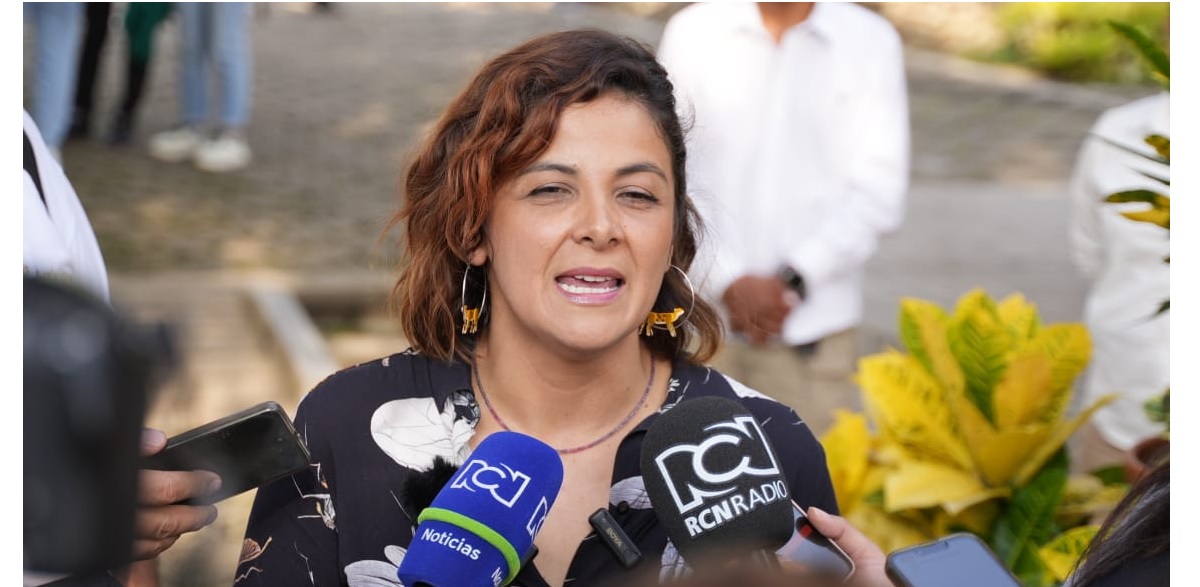 En la imagen se ve a la ministra Jhenifer Mojica que parece estar hablando con un grupo de reporteros o periodistas. Lleva un top estampado en blanco y negro y el pelo corto y rizado. La ministra sostiene un micrófono, lo que sugiere que se dirige a los medios de comunicación o hace una declaración.