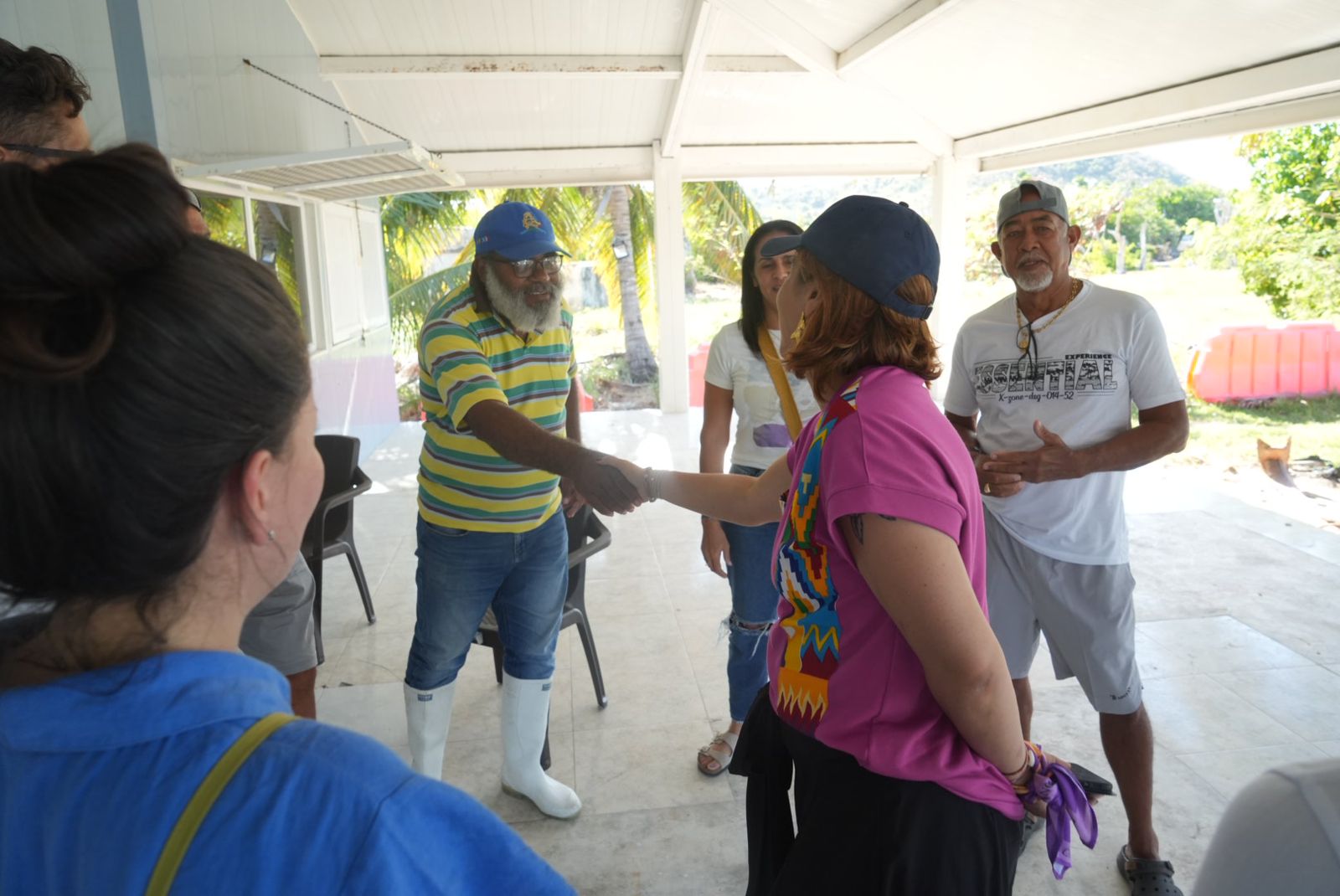 En la imagen se ve a un grupo de personas dándose la mano. La persona de la izquierda parece ser un hombre que lleva una gorra azul y una camisa de rayas de colores, mientras que la persona de la derecha parece ser la ministra Jhenifer Mojica que lleva un top rosa con un diseño de colores en la espalda. Se están dando la mano, lo que suele significar un acuerdo formal, un trato comercial o un saludo amistoso.