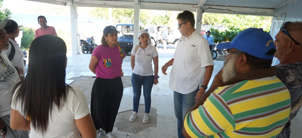 En la imagen, hay un grupo de personas reunidas en lo que parece ser una zona exterior cubierta, posiblemente un patio o un aparcamiento cubierto. Los individuos están conversando, algunos más cerca de la cámara y otros ligeramente distanciados. El ambiente sugiere una reunión informal, posiblemente un acto social o una reunión.