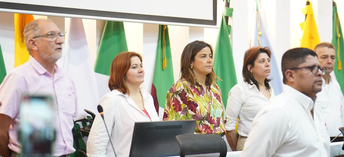 La imagen muestra a un grupo de personas de pie frente a una pared de la que cuelgan varias banderas. El grupo está formado por hombres y mujeres, todos vestidos con camisas blancas. Parece que están reunidos para un acto o una reunión formal, ya que algunos llevan corbata.  Las banderas de la pared son de distintos tamaños y colores, lo que añade diversidad y unidad a la escena.
