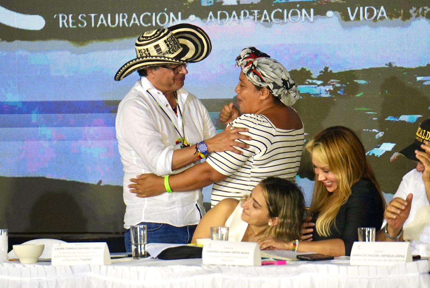 En la imagen hay varias personas reunidas alrededor de una mesa. La persona en primer plano es el presidente Gustavo Petro, que lleva un sombrero tradicional asociado a la cultura Colombiana y abraza a una mujer que lleva un pañuelo en la cabeza.