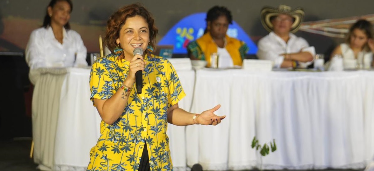 En la imagen se puede ver a la ministra Jhenifer Mojica de pie en primer plano, con un micrófono en la mano y hablando. Parece ser la oradora principal de un acto, posiblemente una conferencia o una reunión. Lleva una camisa de vivos colores, amarillo y azul, que destaca sobre los colores más tenues del fondo.