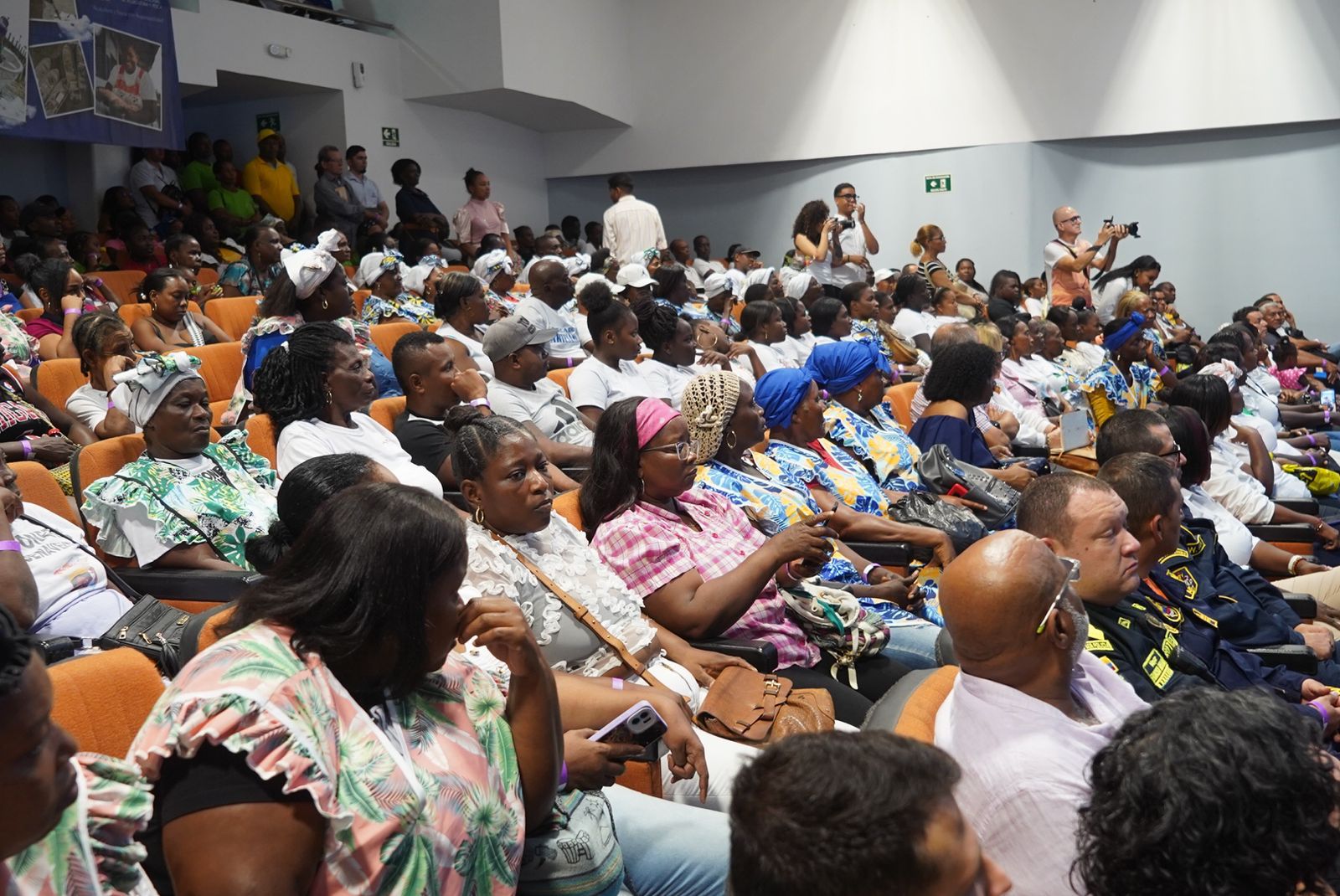 La imagen muestra a un gran grupo de personas sentadas en un auditorio o sala de conferencias. El público parece diverso, con personas de distintas edades y estilos de vestir.