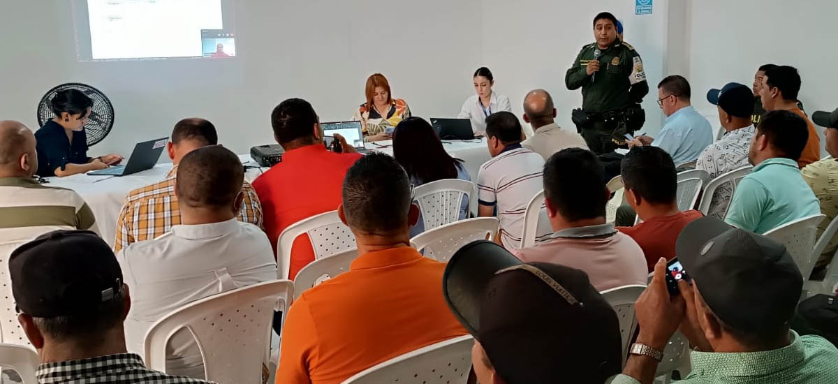 La imagen muestra a un grupo de personas que asisten a una presentación o reunión en una sala. Hay personas sentadas en sillas frente a un frente en el que se está realizando una presentación. El presentador parece estar de pie y hablando, mientras que otros están sentados, algunos con ordenadores portátiles, lo que sugiere que podrían estar tomando notas o siguiendo la presentación.