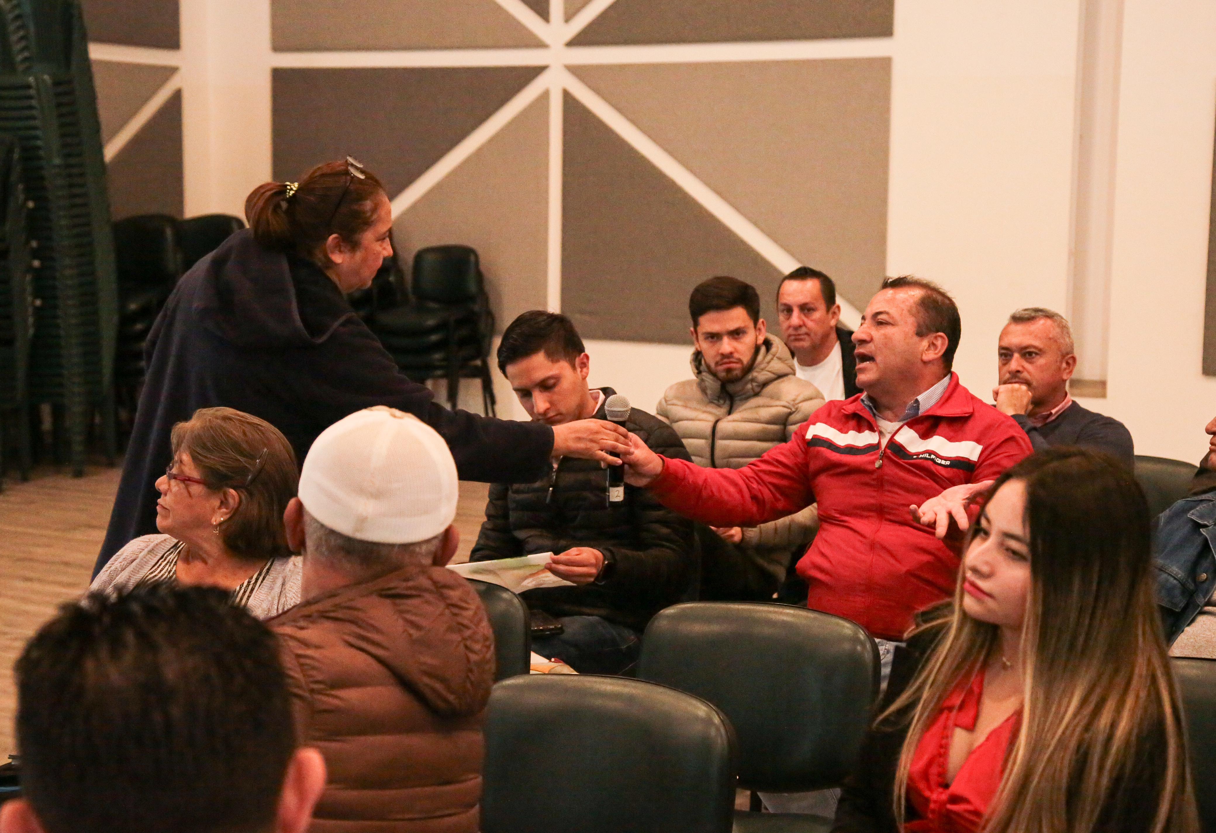 En la imagen se puede apreciar un grupo de personas reunidas en lo que parece ser un evento, la mayoría se encuentran sentados, algunos leyendo documentos y otros participando activamente del evento.