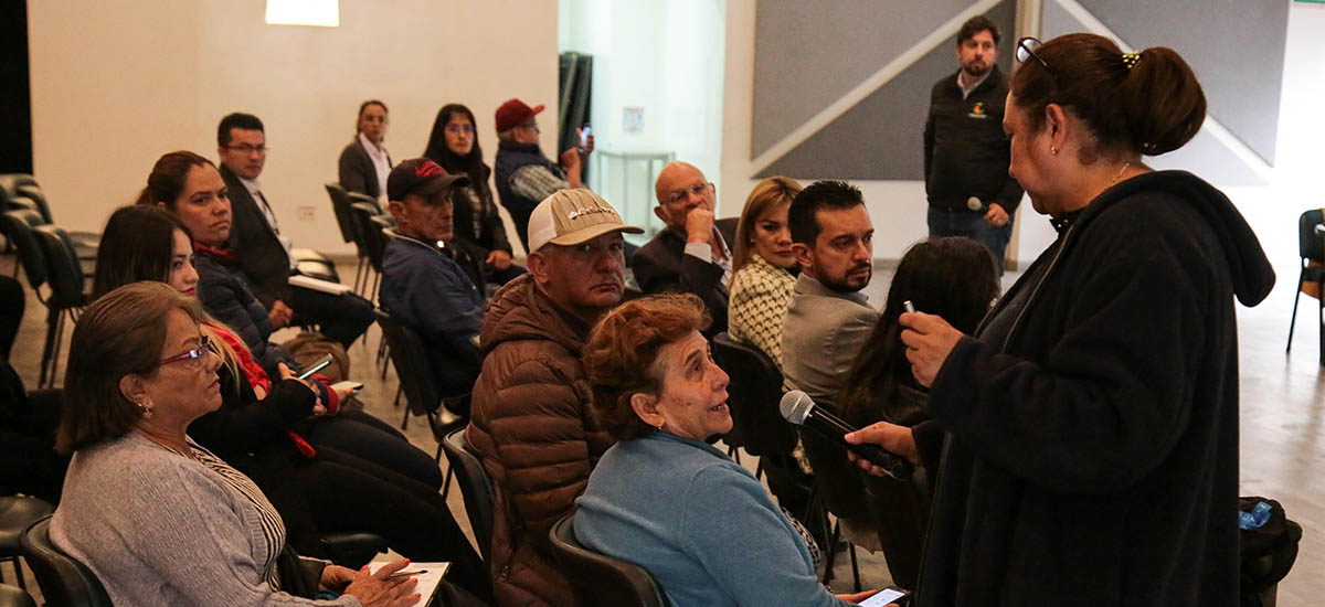 En la imagen se puede apreciar un grupo de personas reunidas en lo que parece ser un evento, la mayoría se encuentran sentados, algunos leyendo documentos y otros participando activamente del evento.