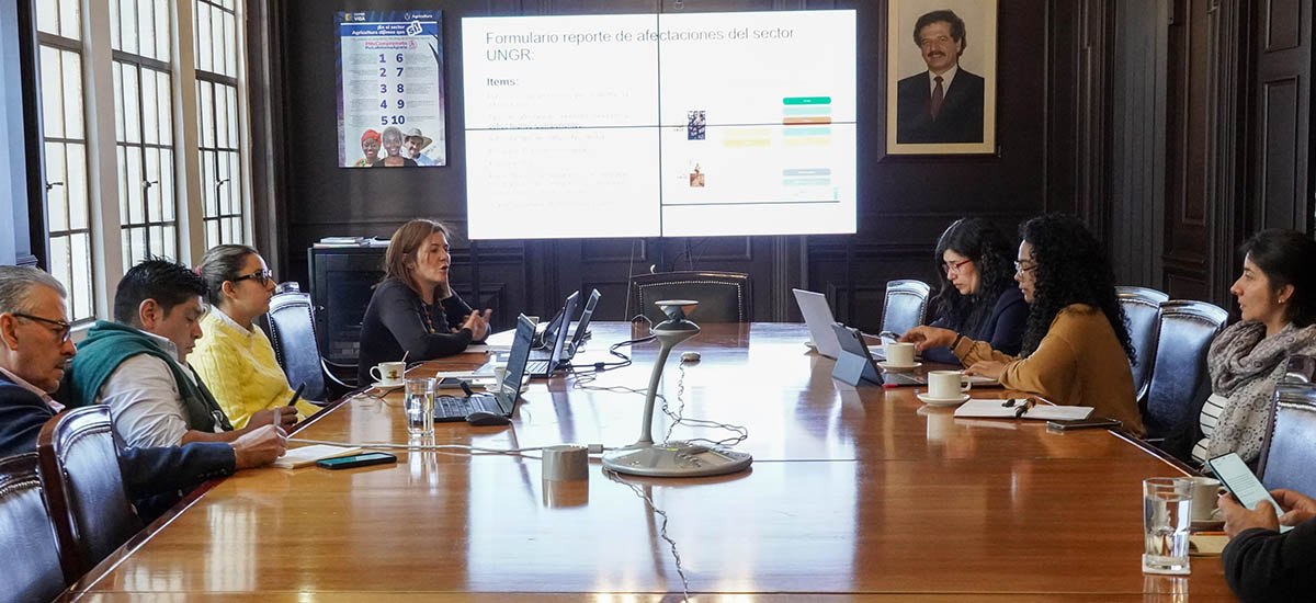 La imagen muestra a un grupo de personas en una sala de reuniones, participando en un debate o una presentación. Están sentados alrededor de una gran mesa, con ordenadores portátiles abiertos frente a ellos, lo que sugiere que están trabajando en un proyecto de colaboración o revisando información.