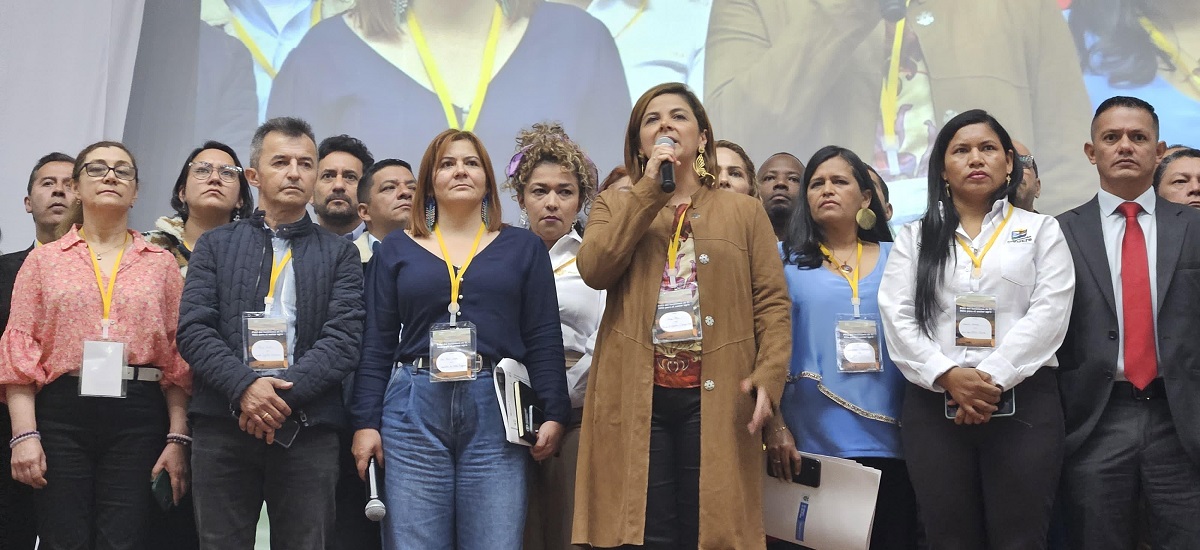 La imagen muestra a un grupo de personas frente a una gran pantalla en la que se proyecta un vídeo o una presentación. La figura central es la ministra Jhenifer Mojica que habla por un micrófono, lo que sugiere que podría estar dirigiendo el acto o haciendo un anuncio.