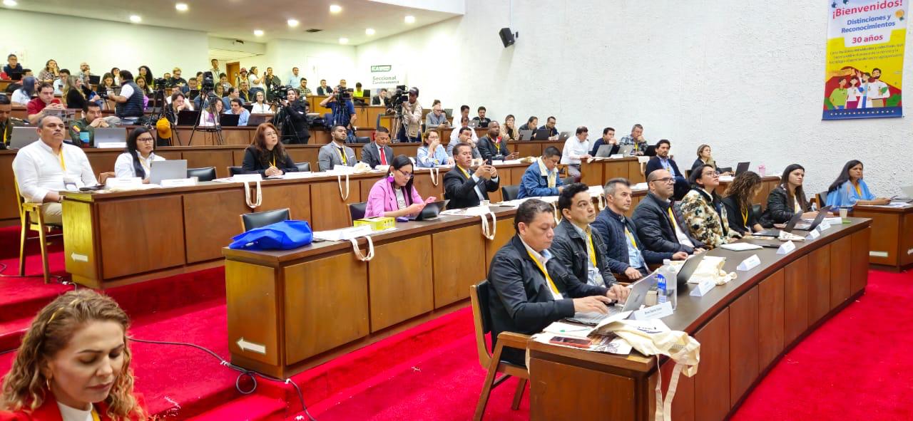 La imagen muestra a un gran grupo de personas sentadas en una sala de conferencias o un auditorio. Los asistentes están dispuestos en filas y algunos están sentados en mesas con ordenadores portátiles y otros materiales. La sala tiene un ambiente formal, con un escenario en la parte delantera donde podría estar sentado un orador o un panel.