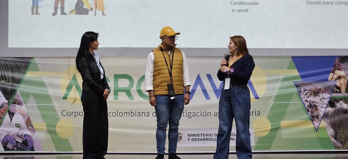 En la imagen aparecen tres personas de pie en un escenario o plataforma, probablemente participando en una presentación o evento. La persona de la izquierda es una mujer la cual lleva un traje negro. La persona del centro es un hombre el cual lleva un chaleco amarillo y una gorra. La persona de la derecha es la Ministra de agricultura Jhenifer Mojica, que sostiene un micrófono y parece estar moderando el acto.
