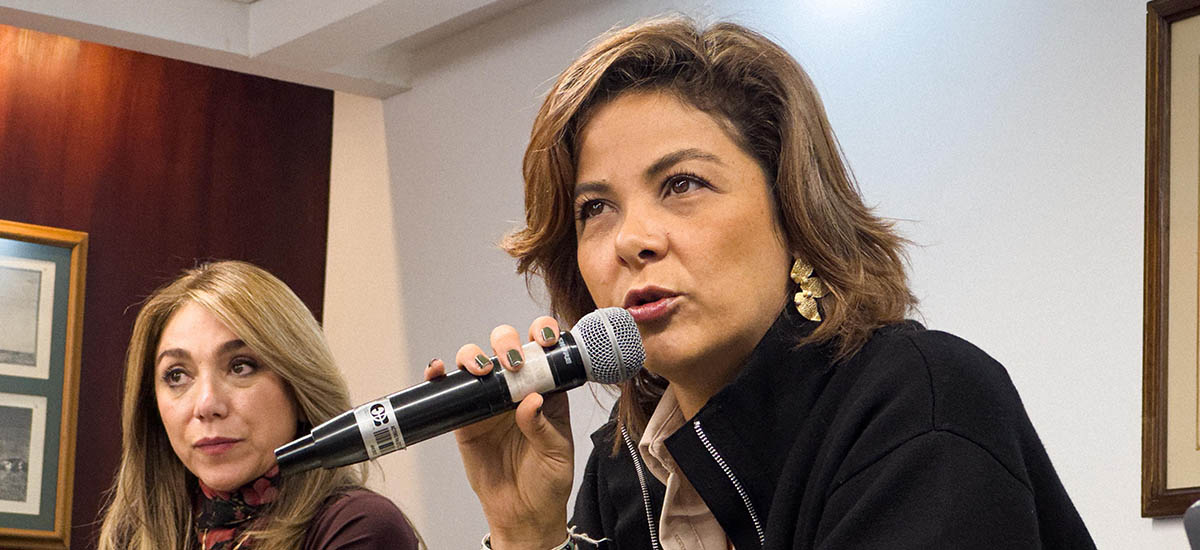 En la imagen se puede ver a la ministra Jhenifer Mojica, ella se encuentra hablando por medio de un micrófono en una sala, atrás de ella se encuentra otra mujer mirando fijamente al receptor.