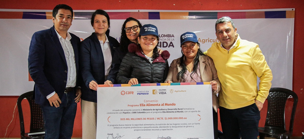 En la imagen aparecen cinco personas posando juntas, probablemente en un acto formal o una reunión profesional. Están de pie delante de un fondo que incluye una pancarta, Dos de los hombres llevan traje y las tres mujeres chaqueta y vestido. Una de las mujeres sostiene un gran documento o certificado, probablemente un premio o reconocimiento relacionado con el evento.