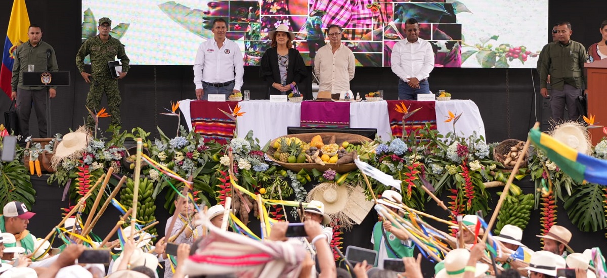 Esta imagen muestra a un grupo de personas de pie detrás de una mesa adornada con una variedad de flores de colores y lo que parece ser un despliegue de frutas. Las personas están vestidas con trajes formales, lo que sugiere que podría tratarse de un evento o ceremonia formal.
