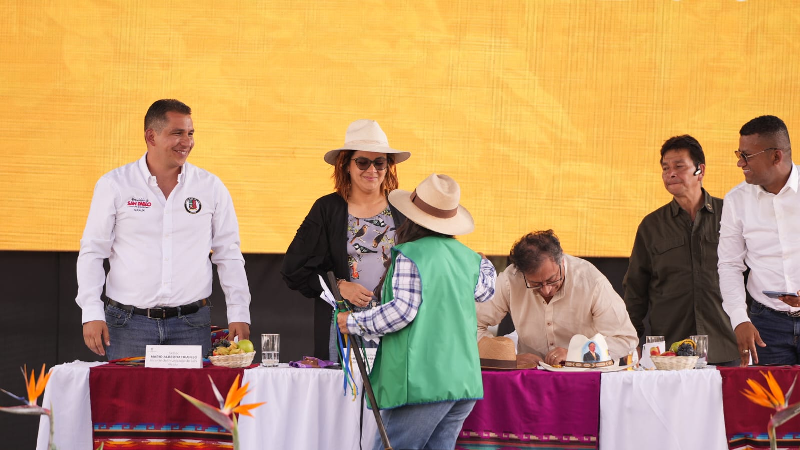 En la imagen, hay un grupo de personas reunidas en torno a una mesa. Parece tratarse de un evento o ceremonia, como indican la vestimenta formal de algunas personas y la presencia de una mesa con lo que parece una tarta y algunos adornos.