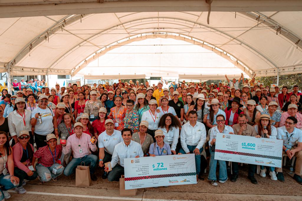 La imagen muestra a un gran grupo de personas reunidas bajo una carpa, posando para una foto. Están de pie frente a un escenario y muchos de ellos llevan sombreros. La multitud está formada por personas de distintas edades y sexos, todas ellas de pie y muy cerca unas de otras.