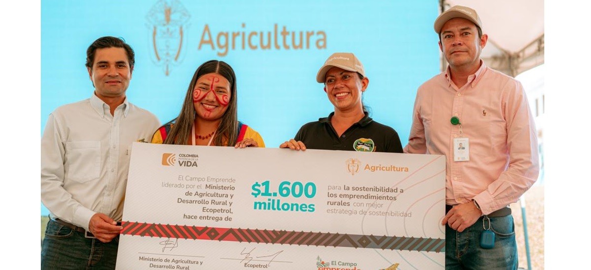 La imagen muestra a un grupo de cuatro personas de pie, posando para una foto. Todos sonríen y parecen felices. Dos de las personas sostienen un gran cheque. El grupo está formado por dos hombres y dos mujeres, todos vestidos con ropa informal. Los dos hombres sostienen el cheque, mientras que las mujeres permanecen de pie a su lado, lo que contribuye al ambiente festivo.