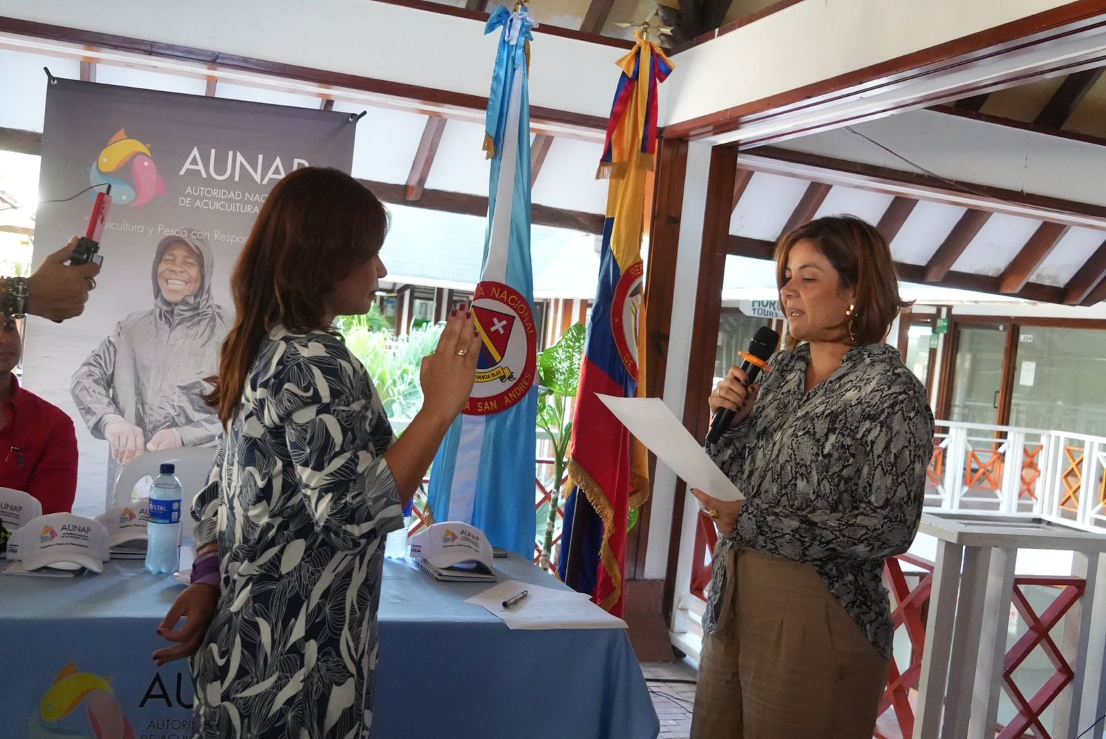 En la imagen, dos mujeres mantienen una conversación. La mujer de la derecha sostiene un micrófono, lo que sugiere que podría estar dirigiendo un debate o moderando un acto. Lleva un top estampado y mira a la otra mujer, que lleva un vestido de flores. La mujer de la izquierda parece escuchar atentamente y sostiene un papel, que podría ser un documento o una lista de preguntas.