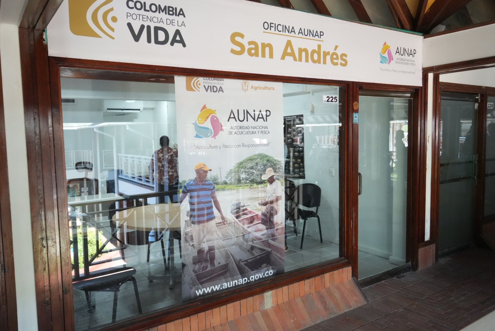 La imagen muestra un escaparate con un cartel en el que se lee "Colombia Potencia de la Vida" y "San Andrés". Debajo del letrero hay un cartel con el logotipo "AUNAP" y el texto "Agricultura y Nacionalidad". El cartel muestra una foto de dos personas, una con una camisa a rayas azules y blancas y la otra con una camisa blanca, de pie frente a un exuberante paisaje verde.