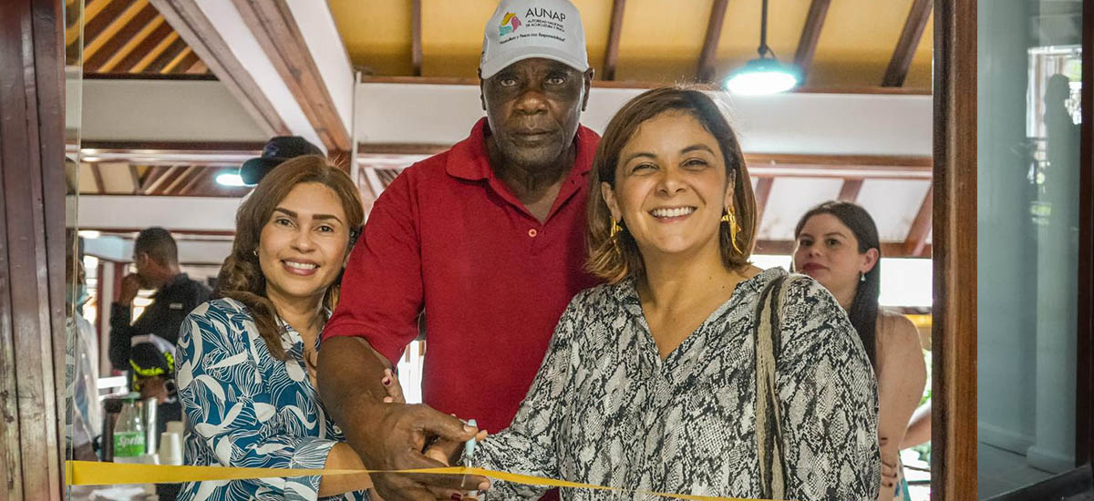 En la imagen aparecen tres personas en un acto formal o semiformal. La persona de la izquierda lleva un top azul estampado y sonríe a la cámara. La persona del centro lleva una camisa roja y una gorra blanca, y está estrechando la mano de la ministra de agricultura Jhenifer Mojica a la derecha, que lleva un top gris con un estampado. La ministra también sonríe y parece estar de buen humor.