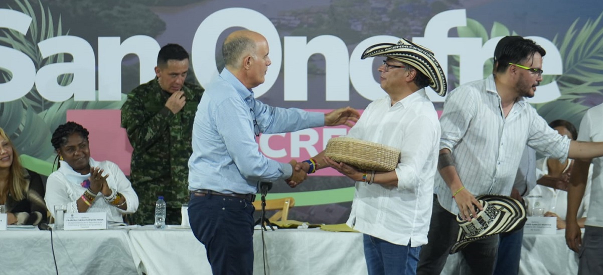En la imagen se puede ver al presidente Gustavo Petro, dando la mano a un ombre y recibiendo lo que parece ser una canasta en un acto publico en el que se ven varias personas más.