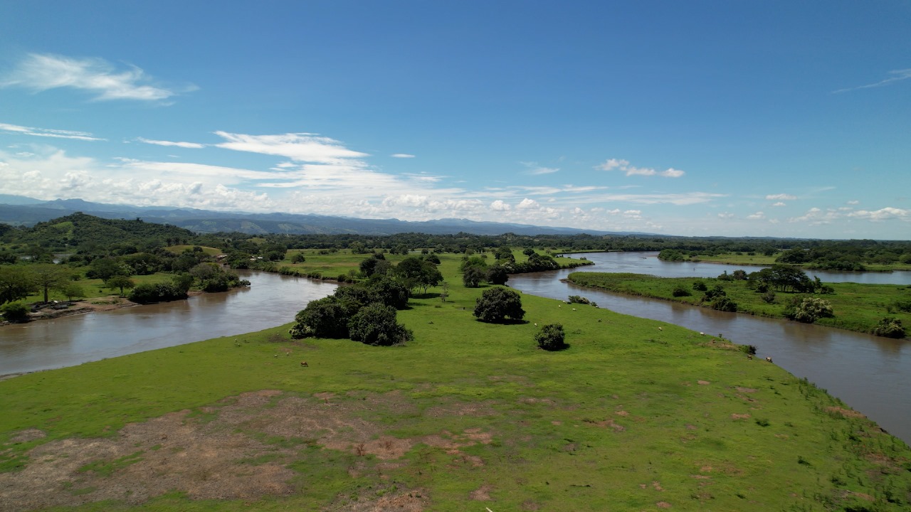 La imagen muestra un gran campo abierto con un exuberante paisaje verde, rodeado por un río en tres de sus lados. El campo está situado en un valle, con montañas visibles al fondo. La escena está captada desde un punto elevado, lo que permite contemplar la zona a vista de pájaro. La combinación del río, las montañas y el extenso campo crea un entorno sereno y pintoresco.
