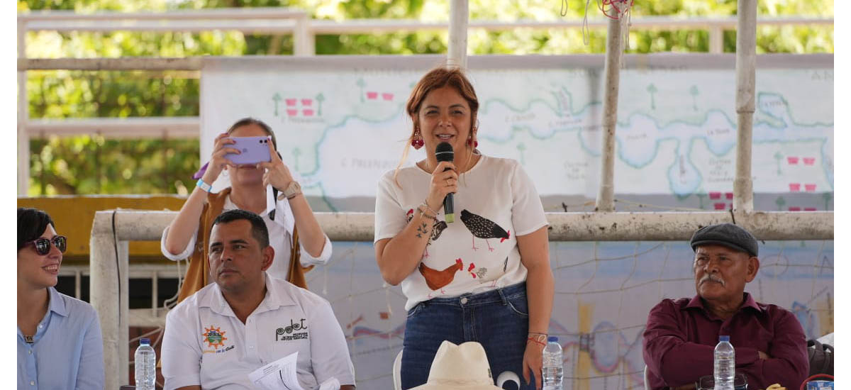La imagen muestra a una mujer de pie en un podio, hablando por un micrófono. Lleva una camisa blanca y parece ser el centro de atención. Hay varias personas entre el público, entre ellas un hombre sentado en una mesa con una botella de agua delante. Otras personas están sentadas y de pie alrededor de la sala, escuchando atentamente el discurso de la mujer.