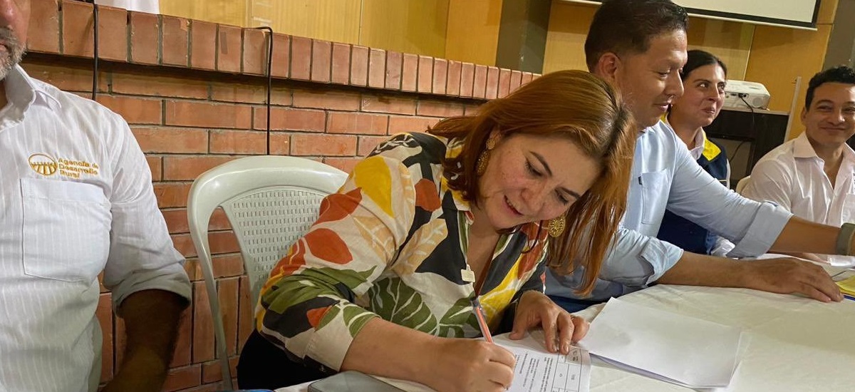 En la imagen aparece la ministra Jhenifer Mojica sentada a una mesa, escribiendo en un papel. Lleva una blusa de colores con motivos florales y parece concentrada en su tarea. Detrás de ella hay varias personas, algunas de las cuales también están sentadas a la mesa, lo que sugiere una reunión o una actividad de grupo. 