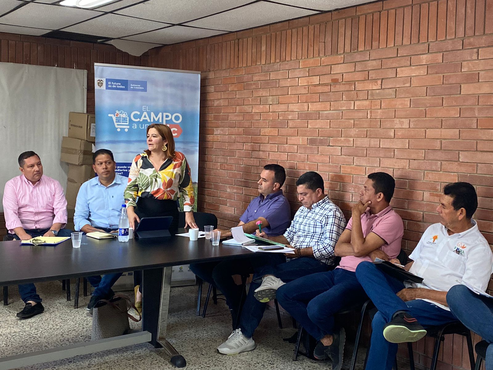 En la imagen, hay un grupo de personas reunidas alrededor de una mesa en lo que parece ser una reunión o una presentación. Se puede ver a la ministra Jhenifer Mojica que está de pie y parece estar hablando o haciendo una presentación al grupo, que está sentado frente a ella. La sala tiene una pared de ladrillo al fondo y hay una pancarta con un texto que parece estar relacionado con el evento o la organización a la que están asociados.a