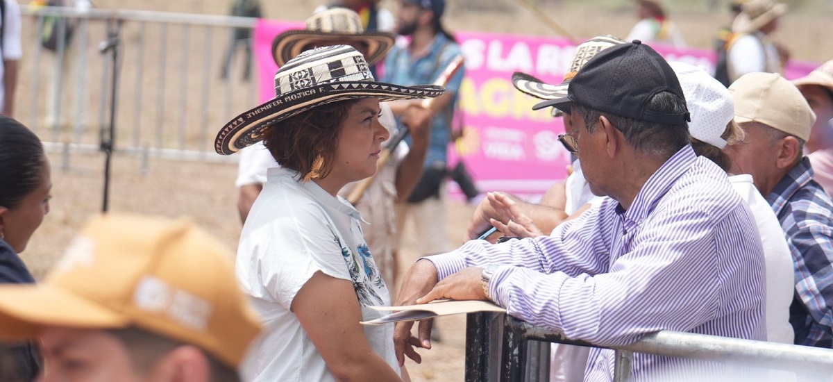 En la imagen se puede observar a la ministra Jhenifer Mojica portando un sombrero tipico de colombia, mientras sostiene una conversación con un hombre que esta inclinada en una valla.