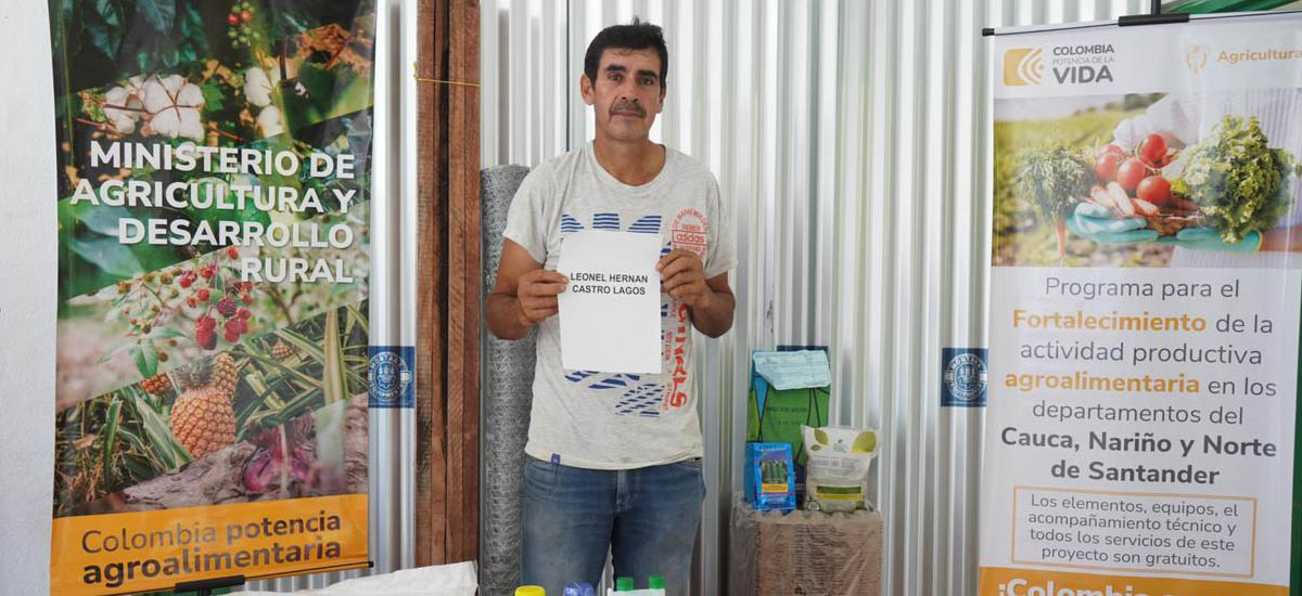 La imagen muestra a un hombre delante de una pancarta con información sobre desarrollo agrícola y rural. Lleva un cartel en el que se lee "LEONARDO CASTILLO", que podría ser su nombre. La pancarta incluye el logotipo del Ministerio de Agricultura y Desarrollo Rural de Colombia y menciona el "Programa para el Fortalecimiento del Sector Agropecuario en los Departamentos de Cauca, Nariño y Norte de Santander".