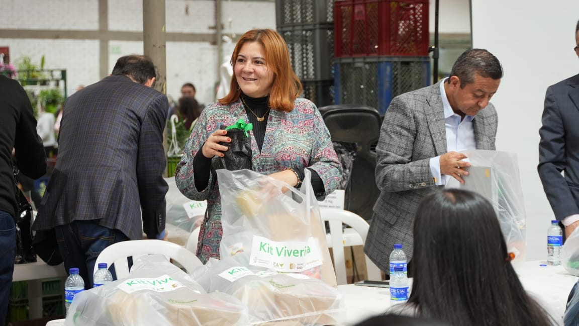 En la imagen hay varias personas reunidas en torno a una mesa con diversos objetos. La Ministra Jhenifer Mojica quien esta en primer plano sostiene una bolsa y parece sonreír, lo que posiblemente indica una interacción o un acontecimiento positivo. En la mesa hay varias bolsas con la etiqueta "KIT VIVERIA", lo que sugiere que podrían contener algún tipo de producto o artículos para su distribución o venta.
