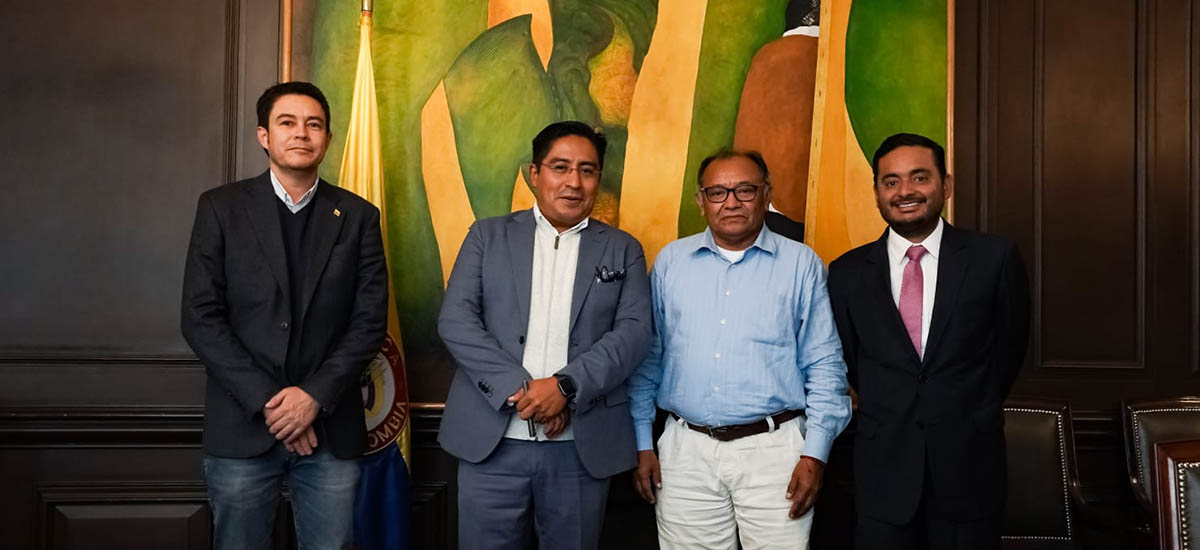 En la imagen se puede ver a cuatro personas posando para una foto, en el fondo se puede observar un cuadro con colores amarillo y verde, también se puede ver una bandera de Colombia a la espalda de uno de los señores.