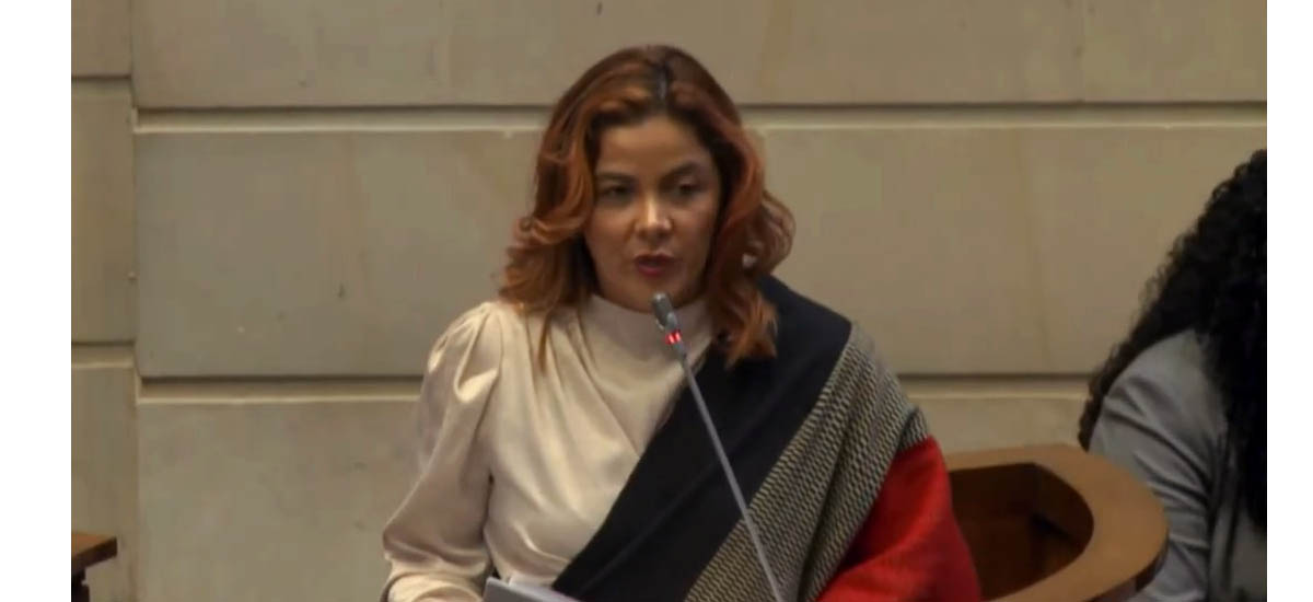 La imagen muestra a una mujer delante de un micrófono, dando un discurso o una presentación. Lleva una camisa blanca y un pañuelo negro, lo que añade un toque de elegancia a su aspecto. La mujer parece ser el centro de atención de la escena, ya que se dirige con confianza a su público.